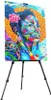 A colorful painting of a woman's face is displayed on a black tripod easel against a white background. Voir les images du produit