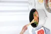 A hand is using a wooden spoon to stir food in a white electric pot. The pot contains shrimp and vegetables. Sliced tomatoes are visible on a plate below the pot. Produktbild-Vorschau