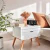 A digital clock with temperature and humidity readings sits on a white nightstand next to a vase of dried flowers and books. Produktbild-Vorschau