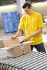 A man in a yellow t-shirt is opening a cardboard box on a conveyor belt in a warehouse. Anteprima dell’immagine prodotto