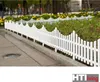 A white picket fence bordering a garden bed with yellow flowers and green plants. Produktbild-Vorschau
