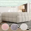 A beige teddy velvet sofa is shown in a living room setting, with three circular close-ups of fabric samples in pink, gray, and white. Produktbild-Vorschau