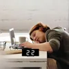 A woman is sleeping at a desk with a laptop and a digital clock. Produktbild-Vorschau