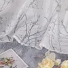 A close-up of a white sheer curtain with a gray branch pattern, draped over a fuzzy gray surface, with a framed picture of flowers and some yellow flowers in the foreground. Produktbild-Vorschau