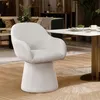 A modern beige armchair is placed next to a marble-topped dining table in a well-lit room with a sofa and plants in the background. Podgląd zdjęcia produktu