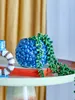 A blue ceramic pot with a succulent plant is placed on top of an orange book, which is on a light green tray. A decorative ring with brown and white stripes is to the left of the pot. Produktbild-Vorschau
