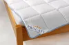 A white quilted mattress topper with a blue trim is shown on a wooden bed frame. Produktbild-Vorschau