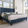 A blue upholstered bed with a tufted headboard is shown in a well-lit room with large windows. Produktbild-Vorschau