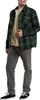 A man is standing and posing in a studio setting, wearing a green and black plaid shirt over a beige t-shirt, and gray pants with black sneakers. Podgląd zdjęcia produktu