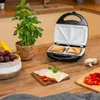 A sandwich maker with two toasted sandwiches inside, placed on a wooden table next to a potted basil plant, a bowl of grapes, and some ingredients for a sandwich. Podgląd zdjęcia produktu