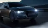 A dark-colored car with its headlights on, driving on a road at night. Produktbild-Vorschau