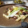 A close-up of a pasta dish with olives and capers on a textured brown plate. Produktbild-Vorschau