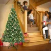 A family is gathered around a Christmas tree in a home setting, with gifts placed around the tree and on the stairs. Produktbild-Vorschau
