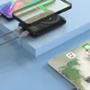 A black power bank with two charging cables connected to it, and a smartphone placed on top of it, suggesting wireless charging. Another tablet is visible in the foreground. Produktbild-Vorschau