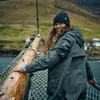 A woman wearing a beanie and jacket stands on a boat with a village in the background. Produktbild-Vorschau