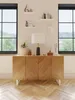A wooden sideboard with decorative items and a lamp on top, placed in a room with large windows overlooking greenery. Náhľad obrázku produktu