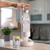 A hand is pressing the top of a Wessper water carbonator on a kitchen counter, with a glass of water and a plant nearby. Produktbild-Vorschau