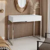 A modern white console table with chrome legs is placed against a slatted wood-paneled wall, with a decorative mirror and dried flowers above it. A chair is visible to the right, and part of a bed to the left. Podgląd zdjęcia produktu