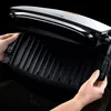 A close-up shot of a person's hands opening a black electric grill. Podgląd zdjęcia produktu