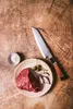 A raw piece of beef steak with some shallots and capers on a plate, next to a small bowl of salt and a knife, all arranged on a wooden surface. Podgląd zdjęcia produktu
