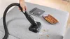 A hand is using a vacuum cleaner attachment to clean crumbs off a gray fabric surface, possibly a sofa or chair. A notebook and a small wooden bowl filled with pretzels are also on the surface. Voir les images du produit