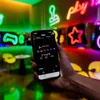 A hand holding a smartphone displaying a colorful interface with various icons and text, set against a backdrop of vibrant neon lights in a room. Produktbild-Vorschau