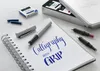 A calligraphy set with pens, ink cartridges, and a notebook is displayed on a white surface. Podgląd zdjęcia produktu