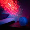 A blue projector is projecting a starry night sky with a crescent moon onto a wall and pillows in a dimly lit room. Produktbild-Vorschau