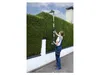 A woman is using a telescopic hedge trimmer to trim a tall hedge. The scene is outdoors with a white wall and a chimney in the background. Produktbild-Vorschau