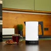 A white electric kettle with a black handle is on a dark countertop, next to a glass mug of tea and a small plate of tea leaves. The background shows a kitchen setting with wooden cabinets and green upper cabinets. Anteprima dell’immagine prodotto
