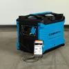 A blue and black EBERTH inverter generator is shown on a concrete floor in front of a garage door. A smartphone is plugged into the generator and displays a website. Produktbild-Vorschau