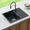 A black granite kitchen sink with a faucet is installed on a countertop, with a blurred background of greenery and a small white mug on a plate. Produktbild-Vorschau