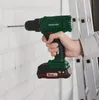 A hand holding a cordless drill, drilling a hole in a white brick wall. A ladder is visible in the background. Produktbild-Vorschau