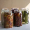 Five glass jars filled with preserved food items, such as pickles, sun-dried tomatoes, and other vegetables, are arranged on a countertop. Produktbild-Vorschau