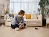 A woman is sitting on the floor in a living room, interacting with a small robot. Náhled obrázku produktu