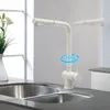 A white kitchen faucet is shown in a modern kitchen setting, positioned over a double sink. Náhled obrázku produktu