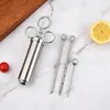 A stainless steel meat injector set with three needles, a cherry tomato, and a lemon slice on a marble-like surface. Voir les images du produit