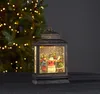 A decorative LED lantern with a winter scene inside, placed on a dark surface in front of a blurred Christmas tree. Podgląd zdjęcia produktu
