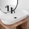 A white oval countertop sink with a black faucet is shown on a wooden vanity. A round mirror is visible in the background. Náhled obrázku produktu