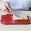 A hand is buttering a slice of bread on a red cutting board. Produktbild-Vorschau
