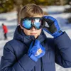 A young boy wearing ski goggles and blue ski gloves adjusts his jacket in a snowy environment. Produktbild-Vorschau
