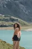A woman stands on a grassy hillside overlooking a turquoise lake and mountains. Produktbild-Vorschau