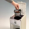 A hand is placing a coffee filter filled with coffee grounds into a coffee maker. Produktbild-Vorschau