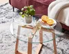 A close-up view of a glass-top coffee table with wooden legs, featuring a plant, a glass of white wine, and sliced oranges on a plate. Anteprima dell’immagine prodotto
