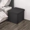 A black storage ottoman is placed next to a bed with patterned pillows. Náhled obrázku produktu