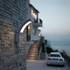 An outdoor wall lamp illuminates a stone wall next to a house with stairs leading up to it. A car is parked in the driveway. Produktbild-Vorschau