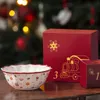 A Christmas-themed advent calendar and bowl are displayed on a wooden surface with a blurred Christmas tree in the background. Produktbild-Vorschau