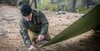 A man is kneeling in a forest setting, using a small axe to secure a green tarp to the ground. Náhled obrázku produktu
