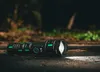 A black and green Vayox flashlight lying on the ground in a forest setting. Produktbild-Vorschau