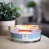 A lit candle with three colored layers in a glass container, placed on a wooden surface next to a potted plant. Náhled obrázku produktu